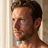 Male model wearing VMEN’S Caffeine + Vitamin C Hydrogel Eye Patches beside a window.